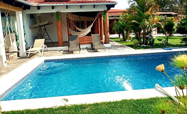 A swimming pool with lounge chairs and a hammock, surrounded by greenery and a building with a tiled roof.