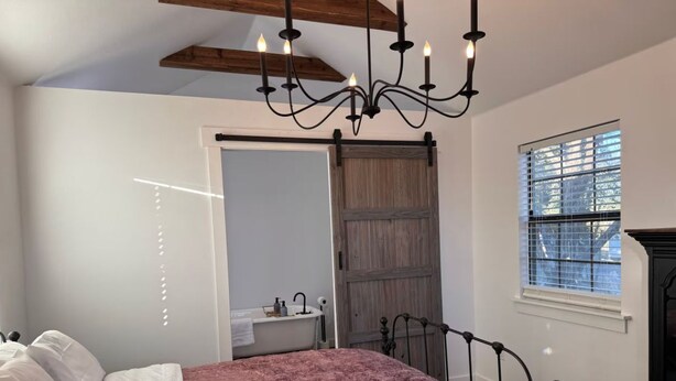 Une chambre à coucher comprenant un lit, un lustre, une fenêtre avec des stores et une porte donnant sur une salle de bain.