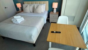 A hotel room with a bed, bedside tables, a desk, a chair, and artwork on the wall.