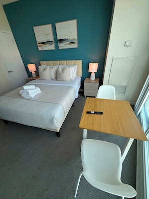A hotel room with a bed, bedside tables, a desk, a chair, and artwork on the wall.