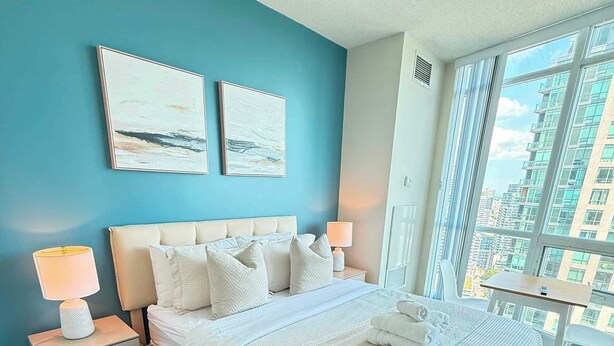 A hotel room with a large bed, two bedside tables, a nightstand, a window with a city view, and two wall-mounted artworks.