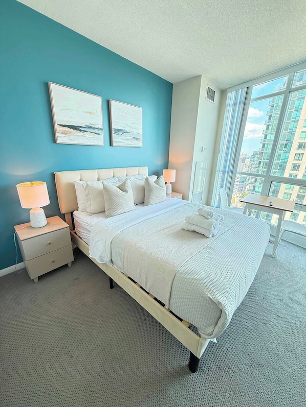 A hotel room with a large bed, two bedside tables, a nightstand, a window with a city view, and two wall-mounted artworks.