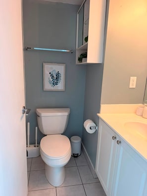 A compact bathroom with a toilet, a small trash bin, a toilet paper holder, and a shelf with a plant.