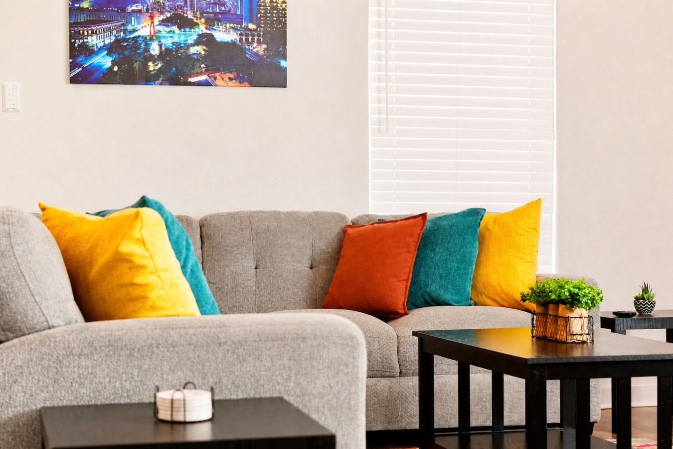 Una sala de estar con un sofá gris, cojines coloridos, una mesita con una planta y un cuadro enmarcado con una vista urbana.