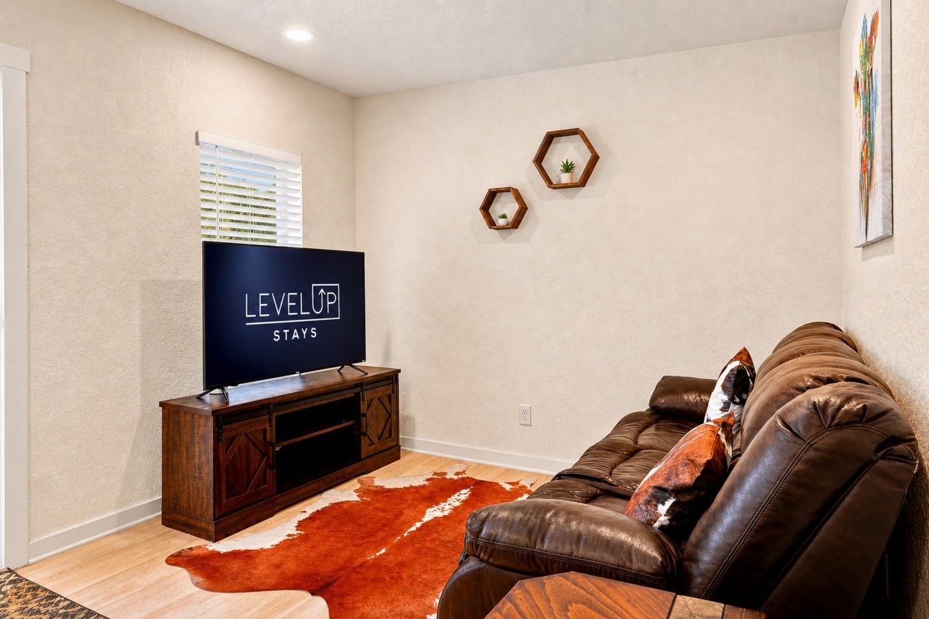 Ein Wohnzimmer mit Fernseher auf einem Holzboden, einem Ledersofa, einem Kuhfellteppich und einem sechseckigen Wandregal mit einer Pflanze.