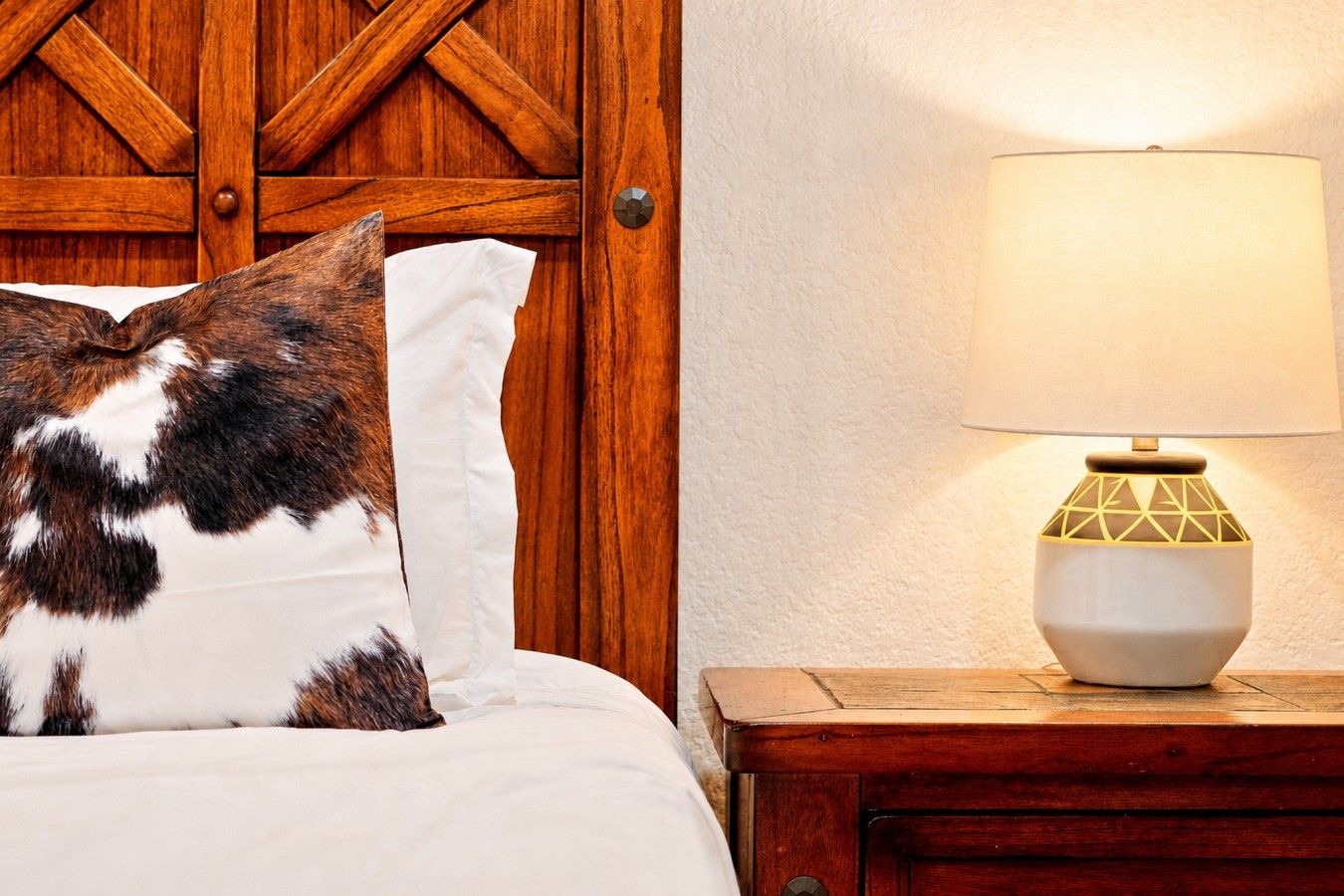 Una cama con un almohadón blanco y uno estampado, una mesita de noche de madera con una lámpara, y un panel de pared de madera.