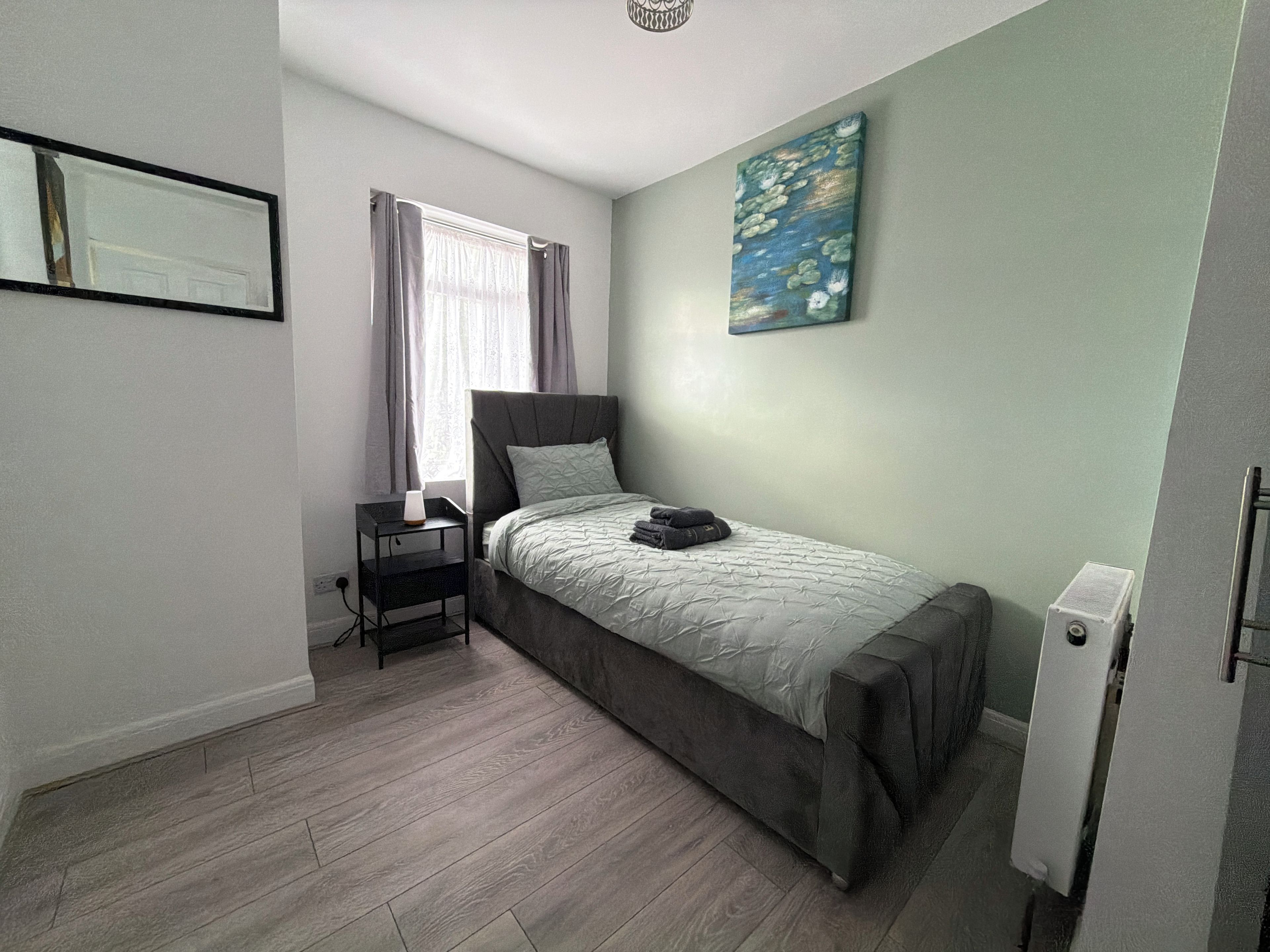A bedroom with a bed, a nightstand, a painting, and a window with curtains.