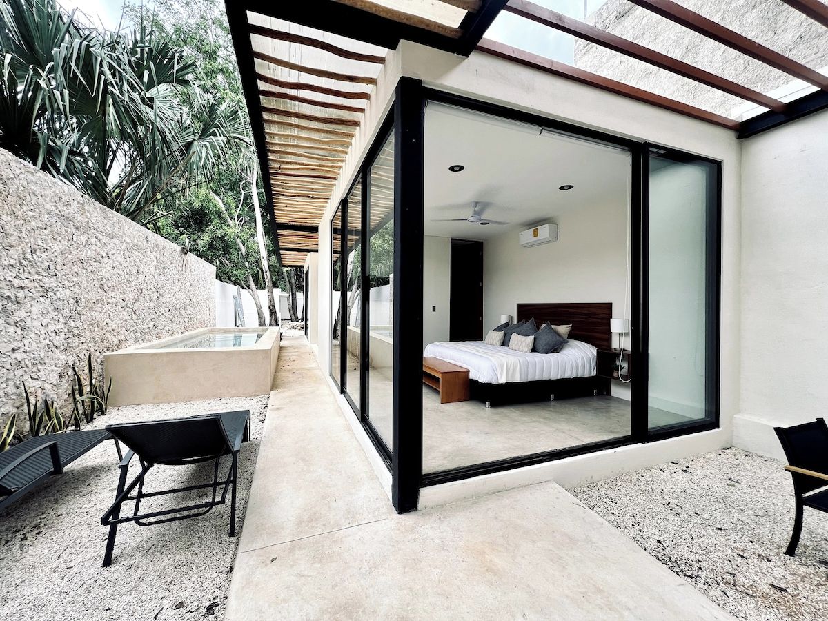 A modern bedroom with a glass door leading to a balcony, a bed with a wooden headboard, and a small seating area with a bench and chairs.
