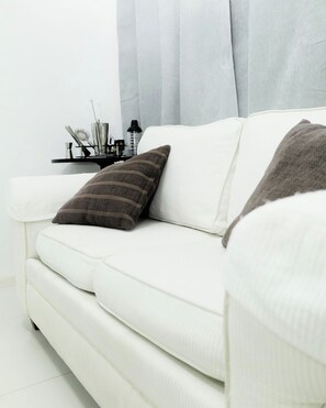 Ein weißes Sofa mit zwei Kissen, ein kleiner Tisch mit diversen Gegenständen und ein Vorhang im Hintergrund.