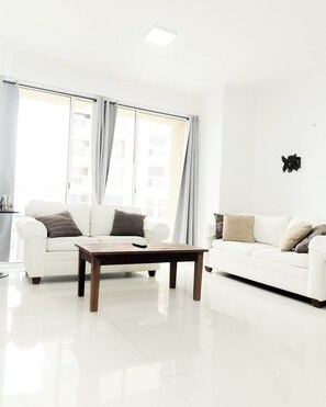 Ein modernes Wohnzimmer mit zwei weißen Sofas, einem Holztisch und großen Fenstern mit Vorhängen.