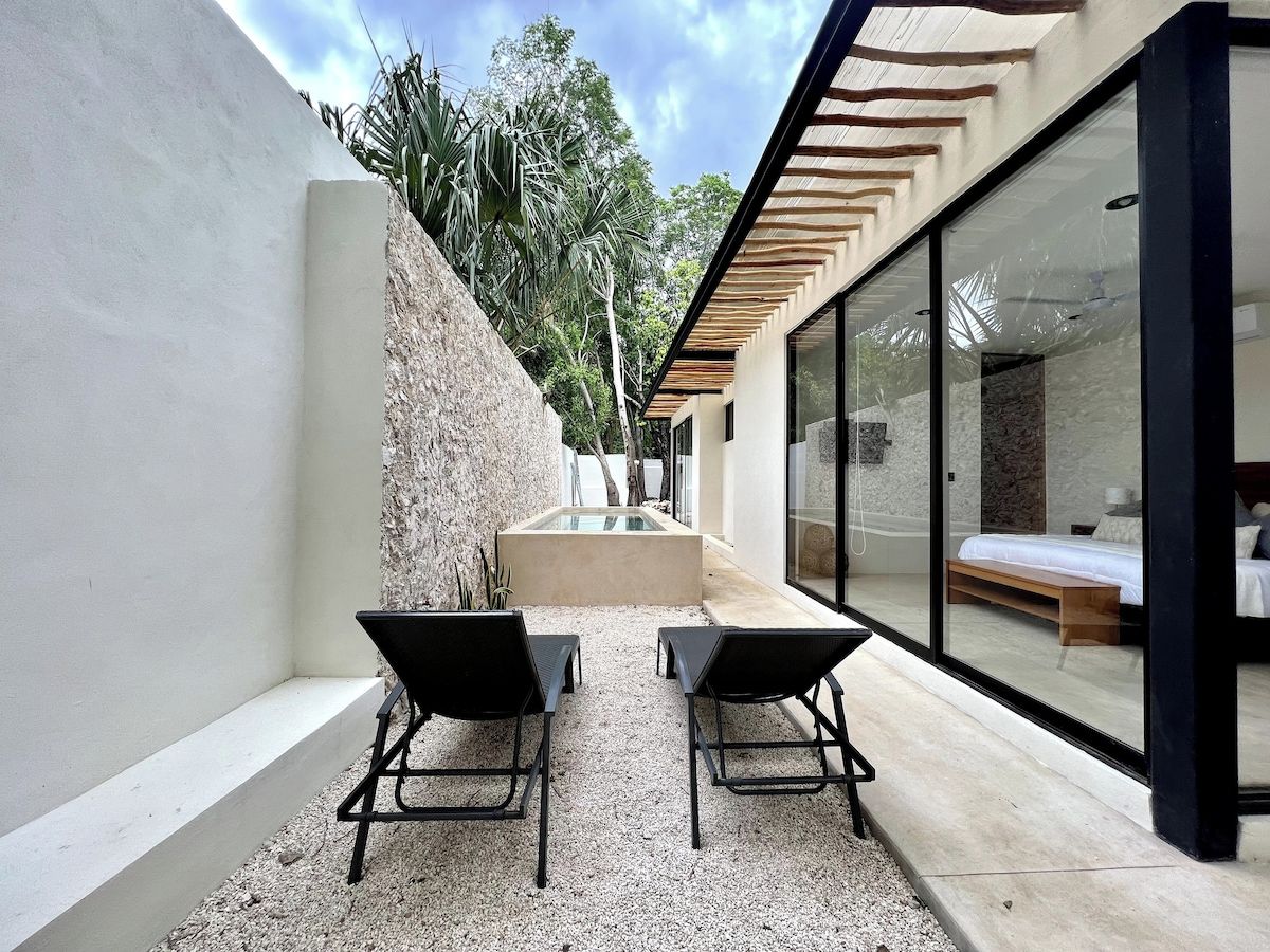 A modern outdoor seating area with two chairs and a small table, a glass-enclosed room with a bed, and a stone wall with a water feature.