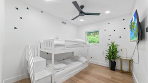 Et værelse med køjeseng, ventilator, fjernsyn, vindue med udsigt til grøn natur og en potteplante.