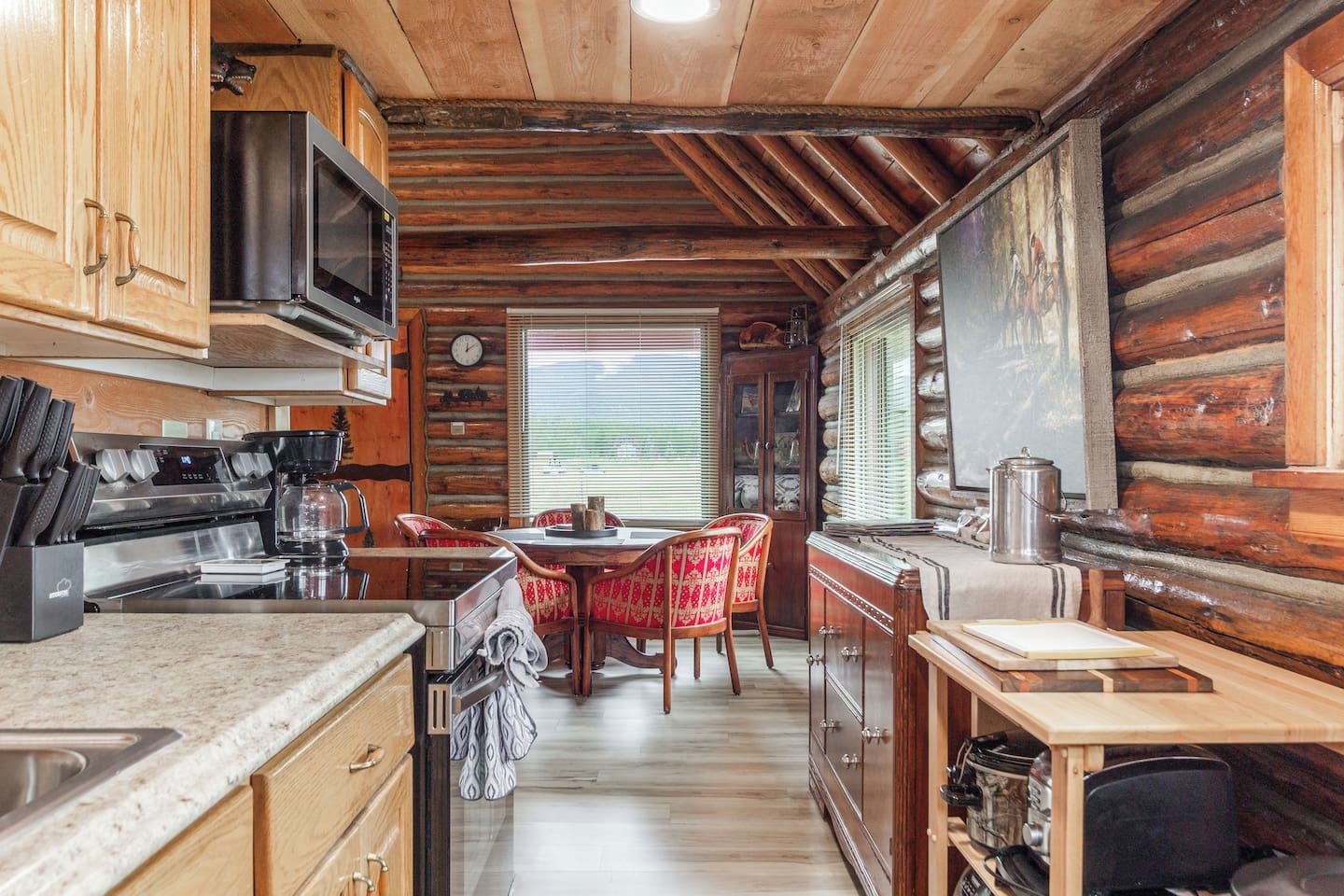 Eine Küchenzeile im Chalet-Stil mit Holzvertäfelung, ein Essbereich mit rot gepolsterten Stühlen und ein großes Fenster mit Jalousien.