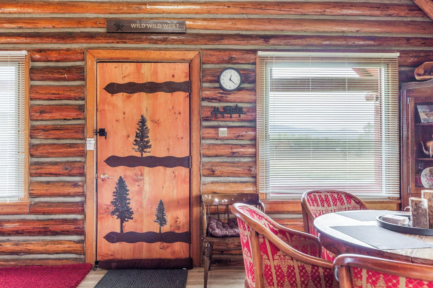 A log cabin interior with a wooden door featuring a tree design, a dining area with a table and chairs, a clock on the wall, and a window with blinds.