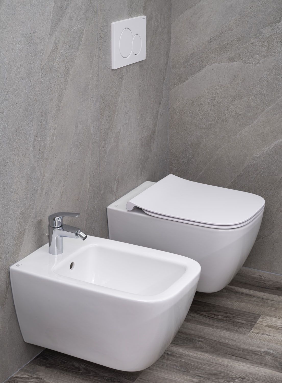 A bathroom with a white bidet, a white toilet, and a wall-mounted flush button.
