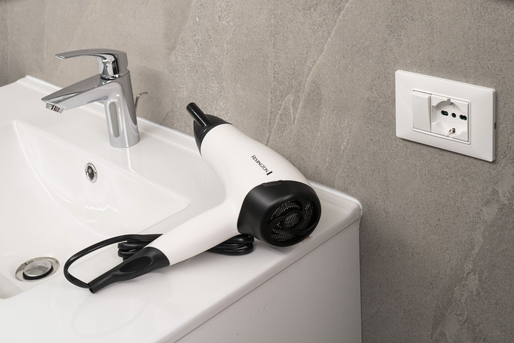 Une salle de bain avec un lavabo blanc, un robinet moderne et un sèche-cheveux branché sur une prise de courant murale.