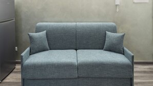 Un sofà gris amb dos coixins a una habitació amb una paret sense decorar i un objecte petit i blanc penjant a la paret.