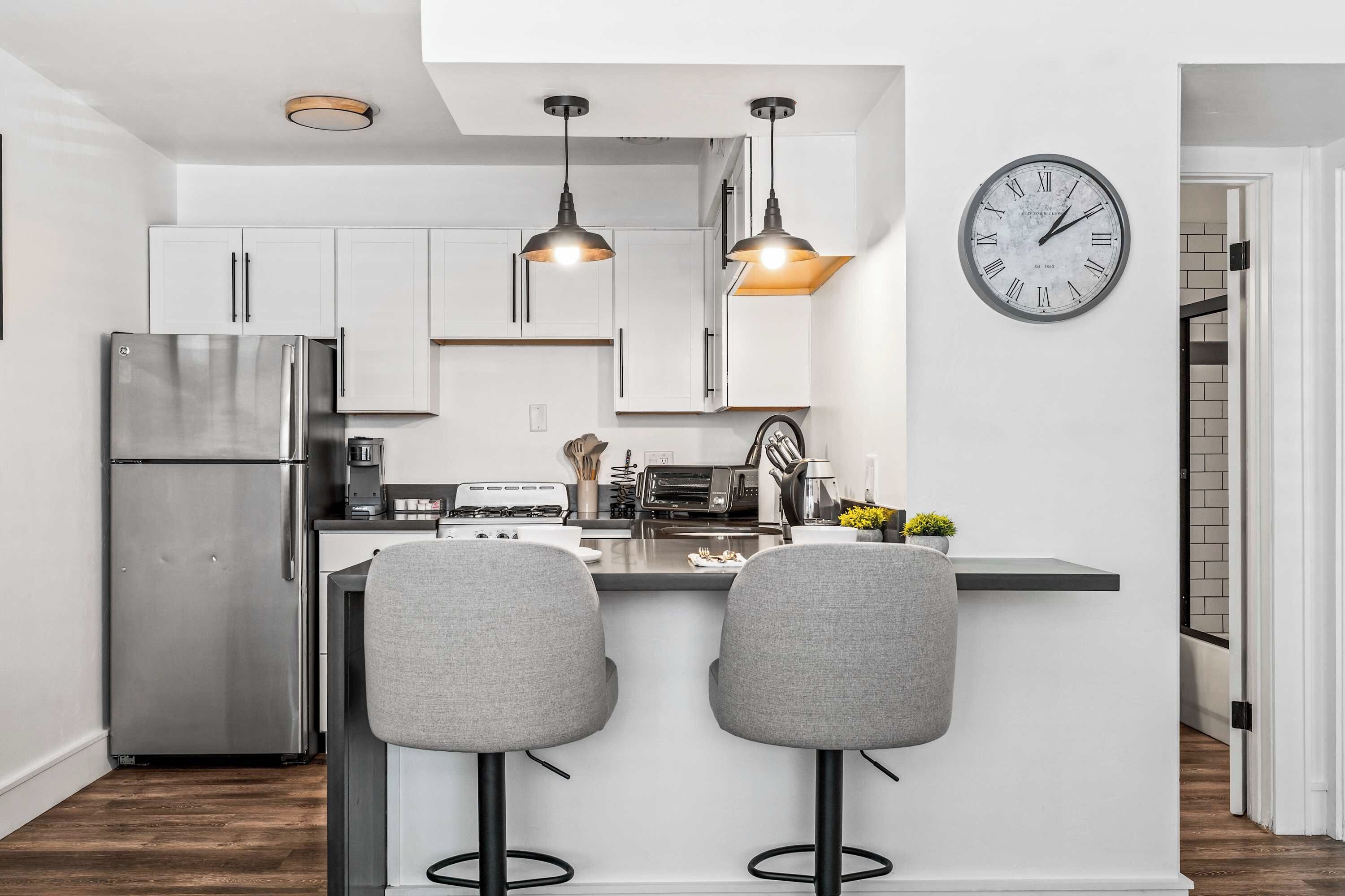 A modern kitchen with a stainless steel refrigerator, two bar stools, pendant lights, and a wall clock.