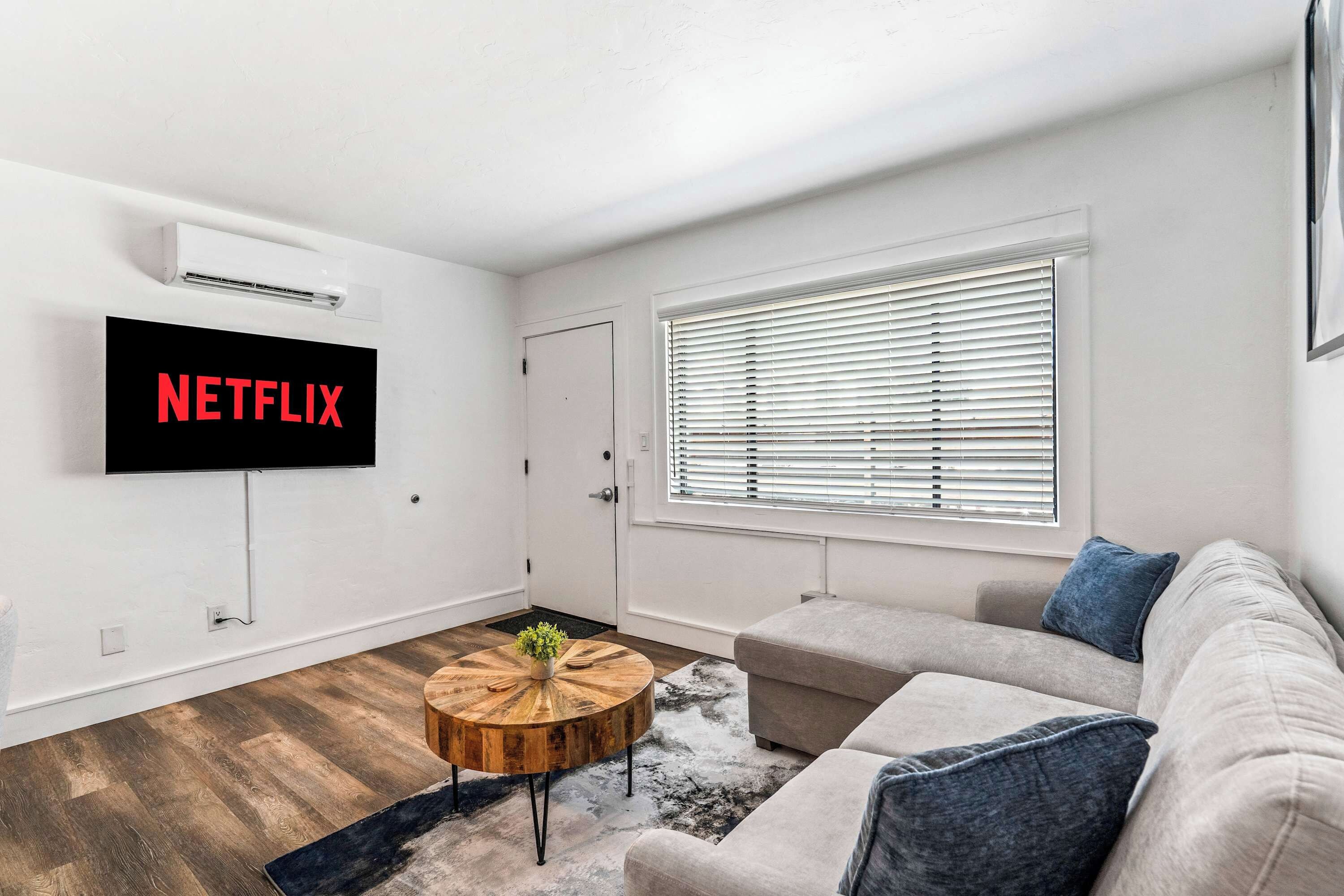 A modern living room with a flat-screen TV mounted on the wall, a round wooden coffee table, a beige sofa with blue cushions, and a window with blinds.
