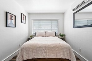 Ein Schlafzimmer mit einem Bett, einem Fenster mit Jalousien, einem Nachttisch mit Pflanze und einem wandmontierten Spiegel.