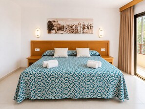 A hotel room with a large bed, a wooden headboard, two bedside lamps, a cityscape painting on the wall, and a view through a window with curtains. - Terrace Apartment for 4 in Pals (Pals)