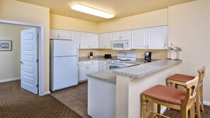 A compact kitchen with white cabinets, a refrigerator, a microwave, a stove, and a coffee maker. There are two wooden bar stools with red cushions.