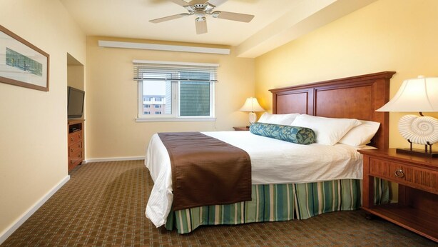 A hotel room with a bed, a nightstand, a lamp, a ceiling fan, a window with blinds, and a television on a dresser. - Long Beach WA Fireplace 2BR Queen Suite – WorldMark Long Beach (Long Beach)