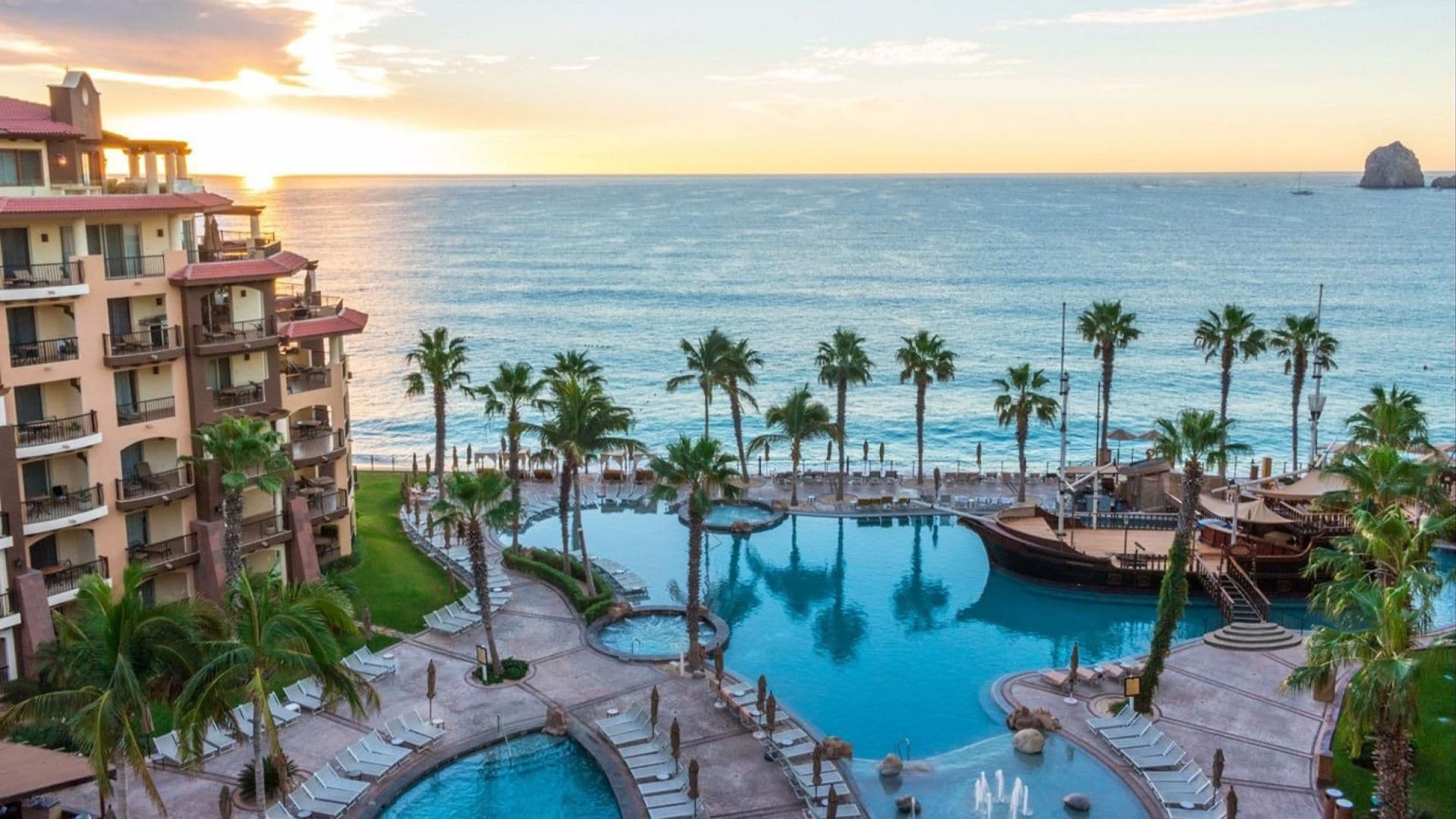 A resort with a large pool area, palm trees, and a view of the ocean at sunset.