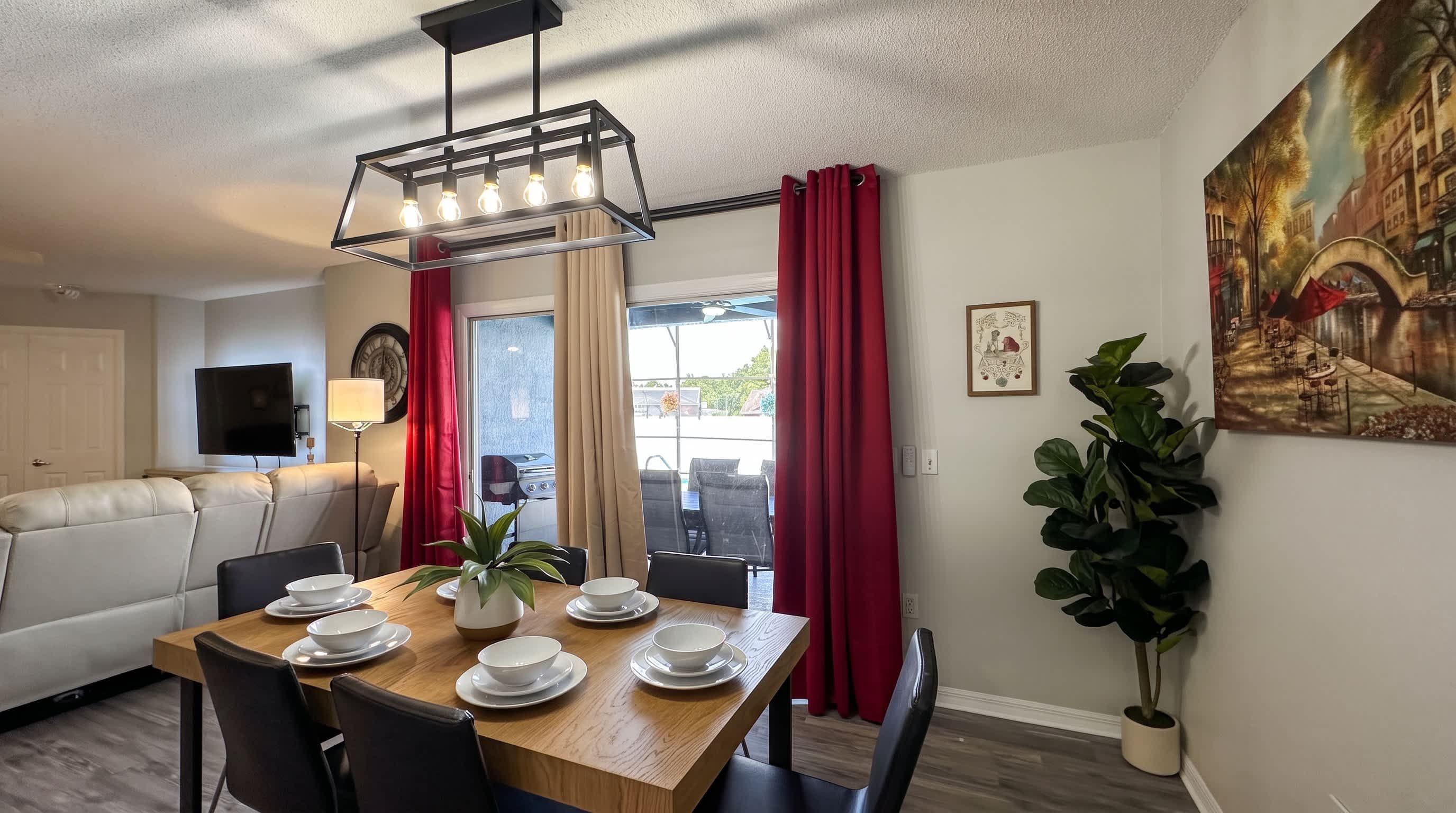 A dining area with a wooden table set for four, a sofa, a television, and a painting on the wall.