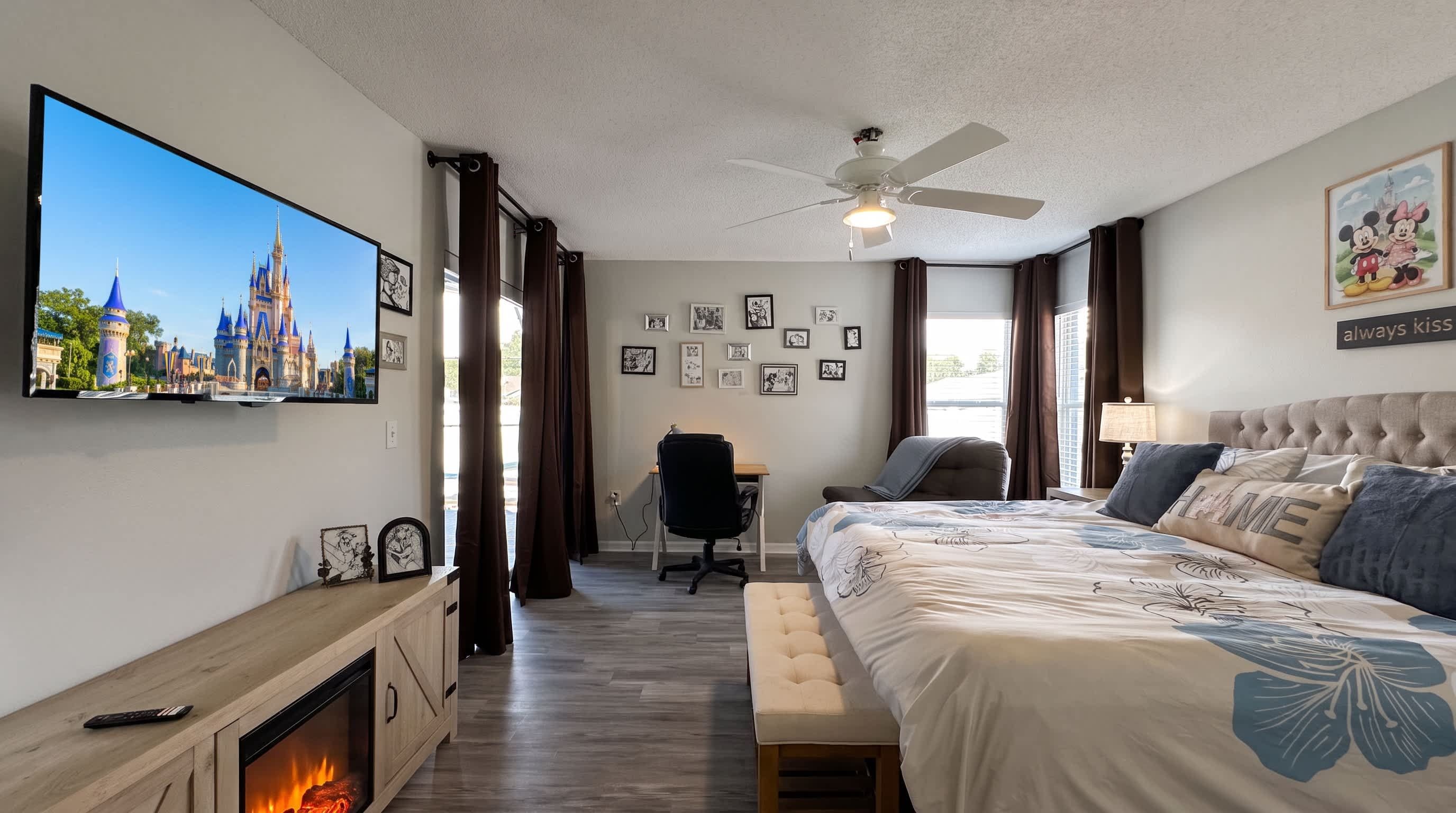 Een hotelkamer met een grote flatscreen-tv, een bed met een gestoffeerd hoofdeinde, een bureau met stoel, een open haard en een plafondventilator.