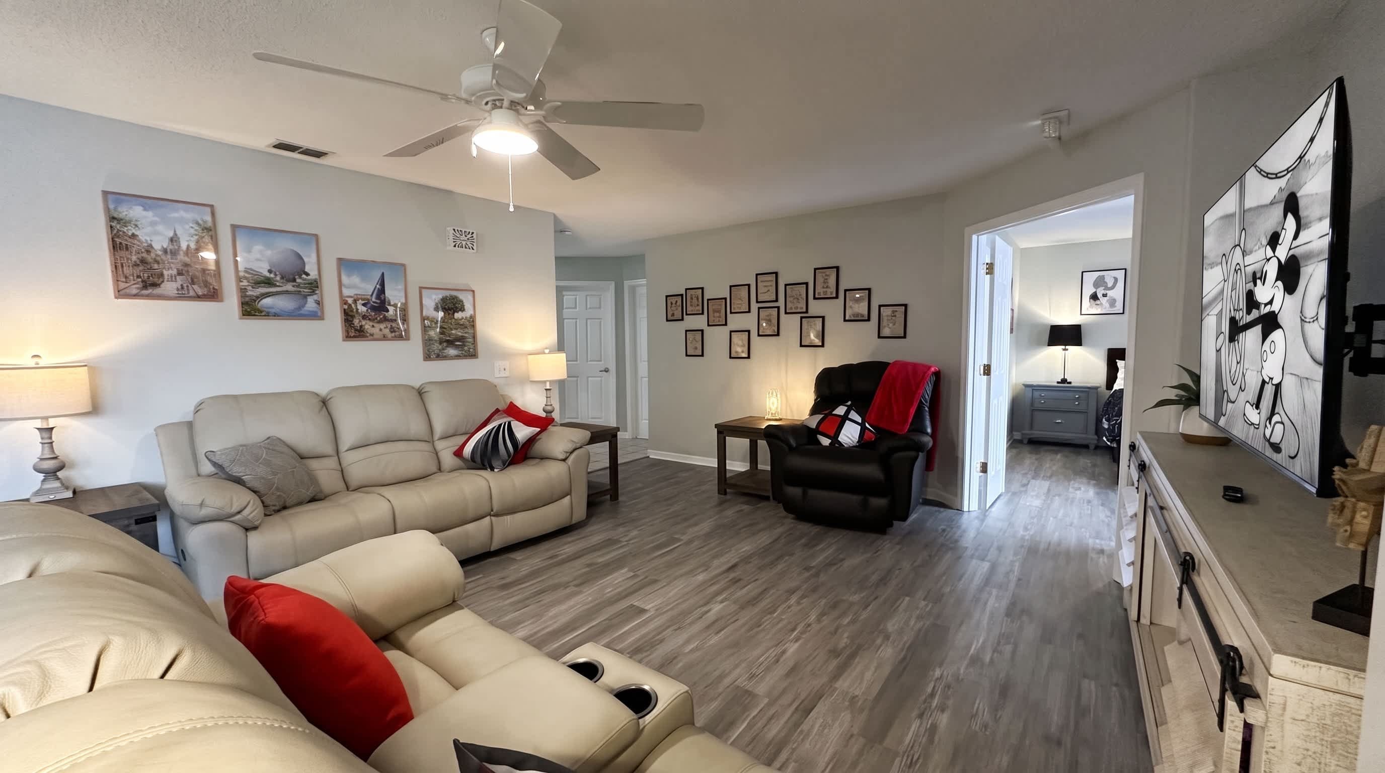 Un salon avec un canapé d’angle beige, un fauteuil noir, un ventilateur de plafond et un téléviseur diffusant un dessin animé.