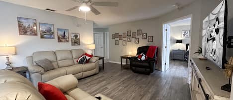 Un salon avec un canapé d’angle beige, un fauteuil noir, un ventilateur de plafond et un téléviseur diffusant un dessin animé.