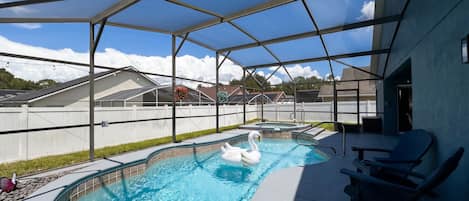 An outdoor swimming pool with a white swan float, surrounded by a covered patio area with chairs.