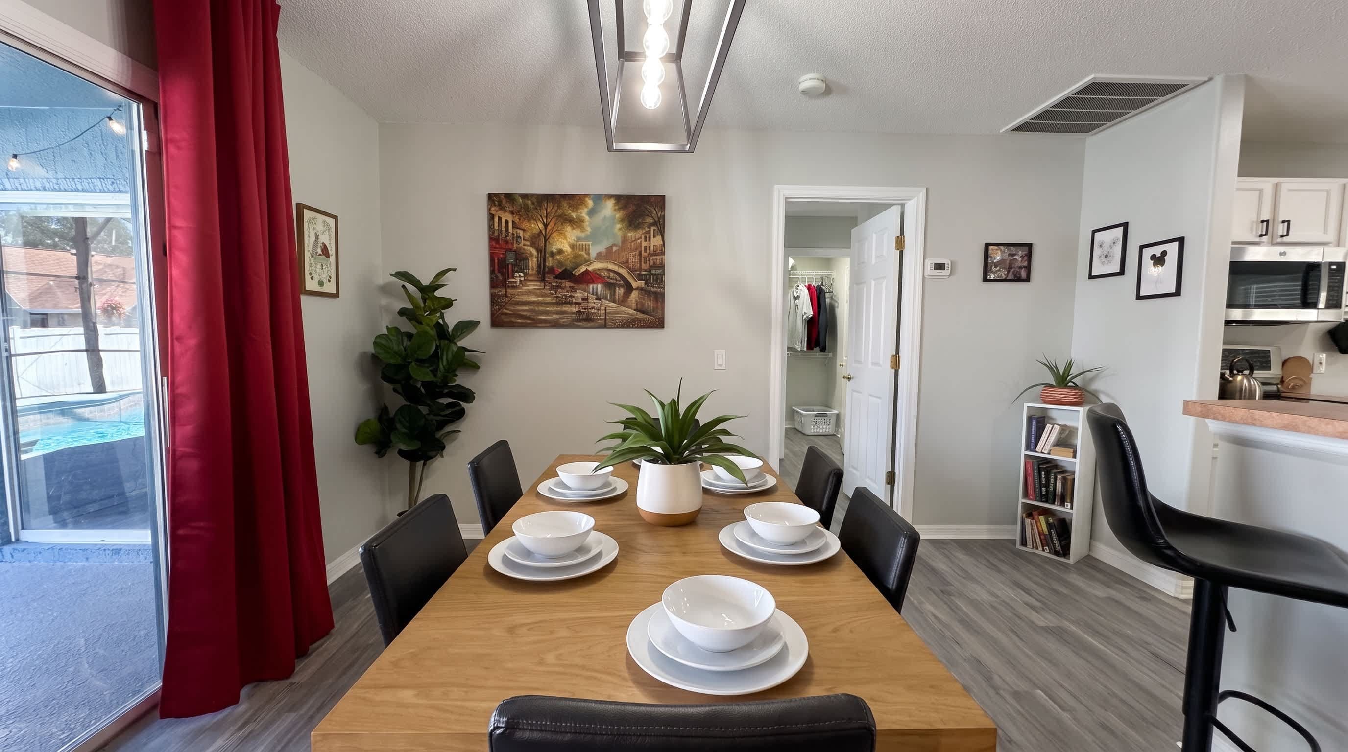A dining area with a wooden table set for four, surrounded by chairs, with a potted plant and artwork on the wall.
