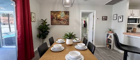 Un comedor con una mesa de madera dispuesta para cuatro personas, rodeada de sillas, una planta en maceta y una obra de arte en la pared.