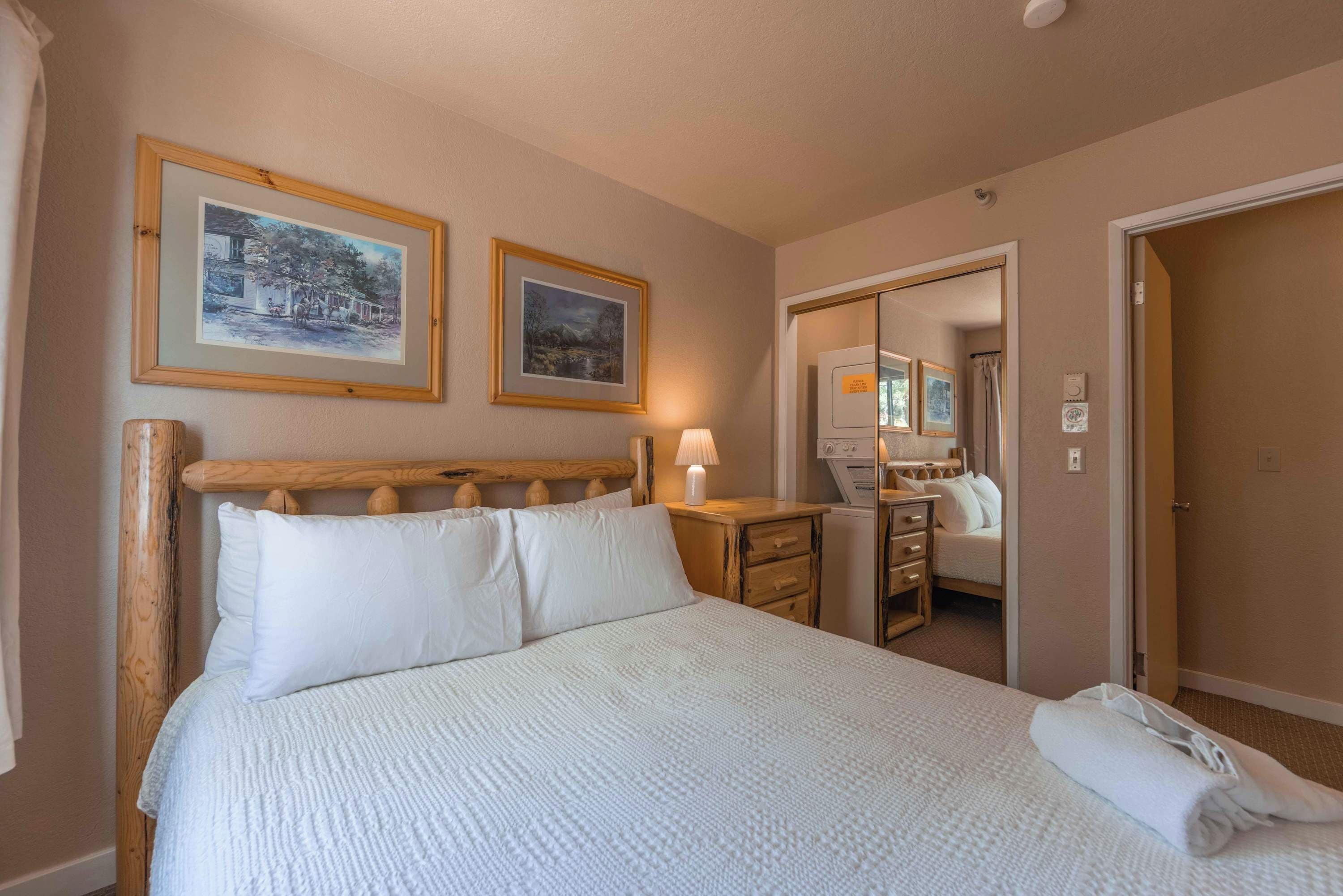Quarto de hotel com uma cama grande, duas fotografias emolduradas na parede, uma mesa de cabeceira com abajur e um espelho.