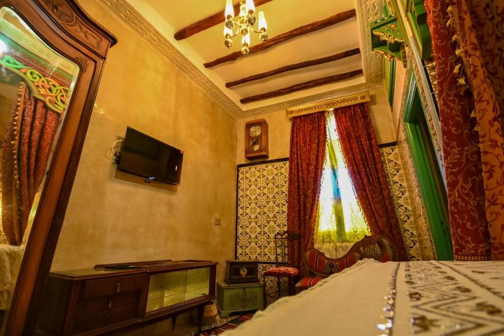 Une chambre traditionnelle avec un lit, une télévision, une armoire en bois et une fenêtre agrémentée de rideaux à motifs.