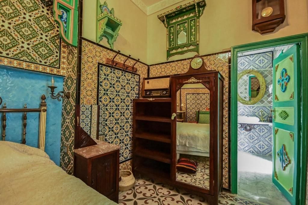 Una habitación con una cama, un estante de madera, un espejo y una puerta con un diseño decorativo.