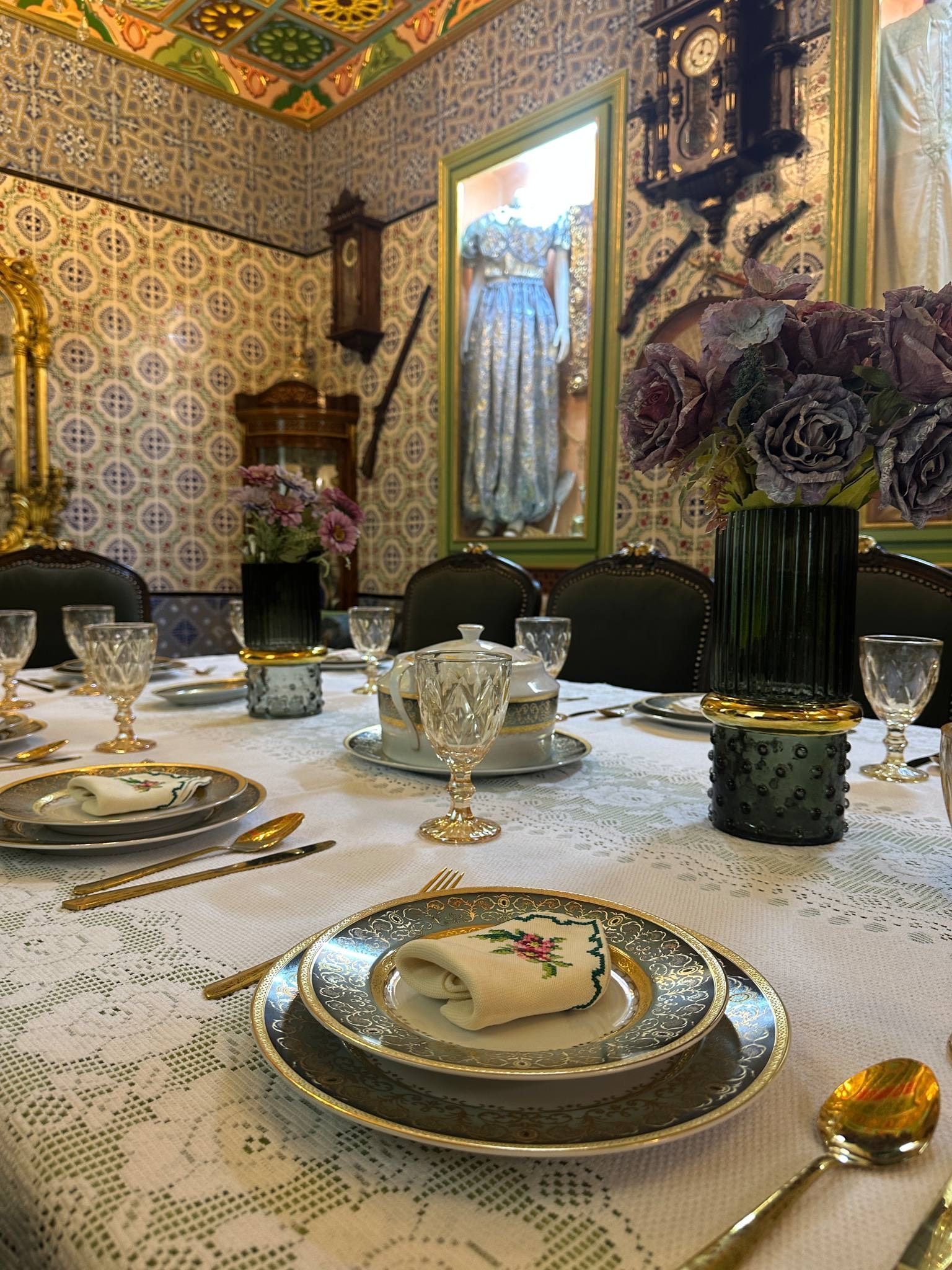 Et spisebord dækket med fint porcelæn, sølvtøj og en vase med tørrede blomster.