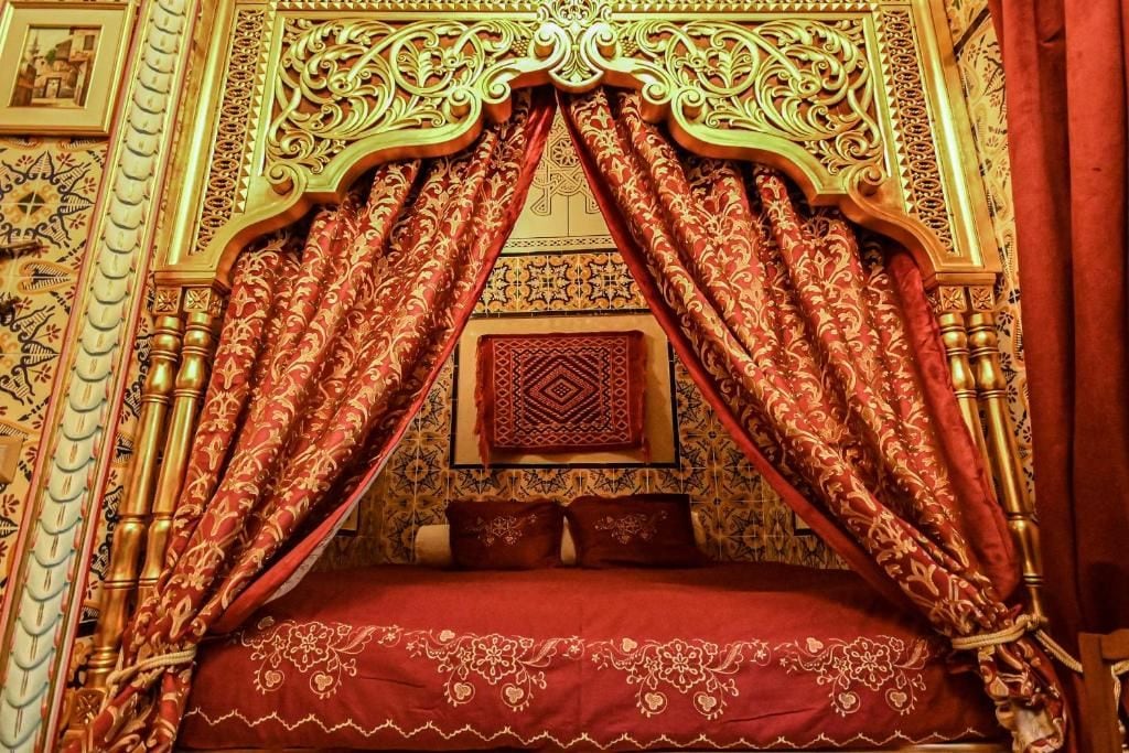 A bed with ornate headboard and red curtains, a patterned wall, and a decorative pillow.