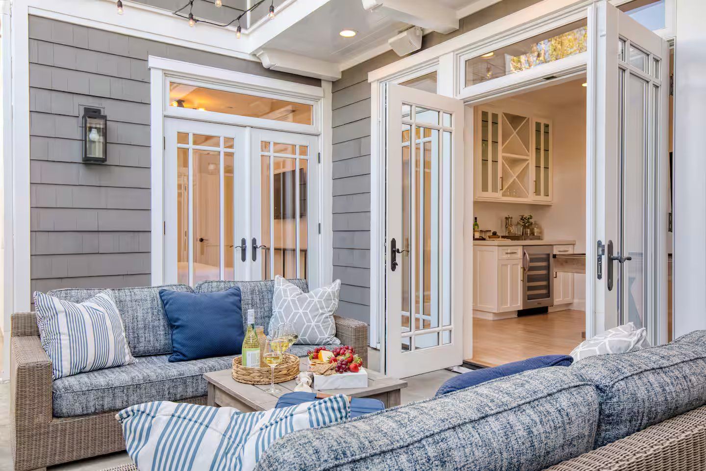 A covered outdoor seating area with a sofa and cushions, a table with a bottle and fruit, and a view into the house through open doors.