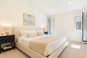 Une chambre à coucher comprenant un lit, des tables de chevet, une commode, une lampe fixée au mur, une fenêtre avec des rideaux et un luminaire au plafond.