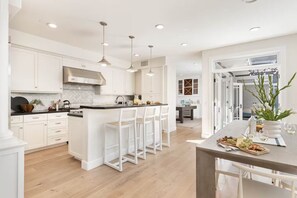 A modern kitchen with white cabinetry, a central island with bar stools, pendant lighting, and a dining area with a table set for a meal. - Mins To Newport Beach, Pier, Water-Front Dining (Newport Beach)