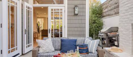 An outdoor patio area with a wicker sofa, a table with wine glasses and a bottle, and a barbecue grill.