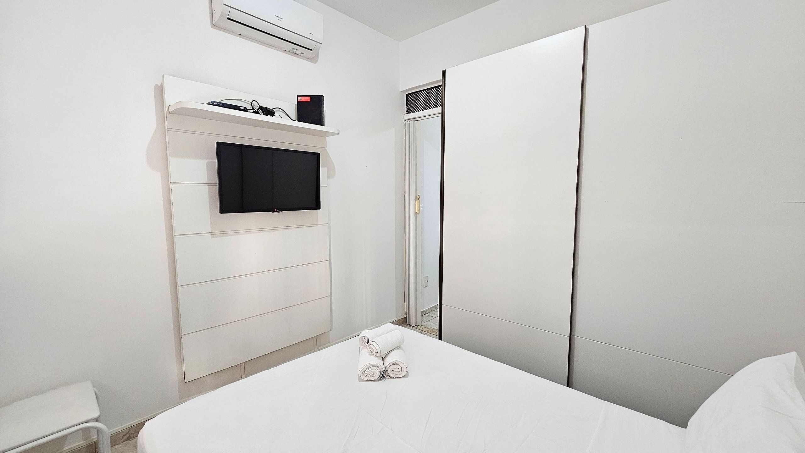 A hotel room with a bed, a TV mounted on a wall, a wardrobe, and an air conditioning unit.