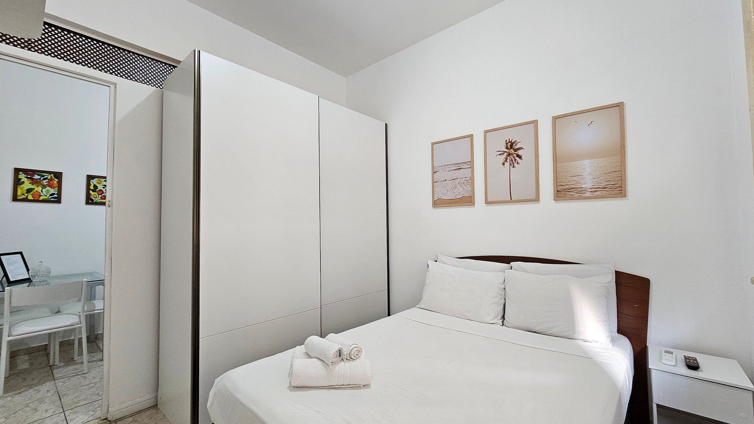 A hotel room with a bed, a nightstand, a desk with a chair, a wardrobe, and three framed pictures on the wall.