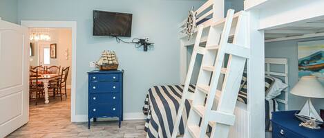 A bedroom with a bunk bed, a desk with a chair, a dresser, and a television on the wall.