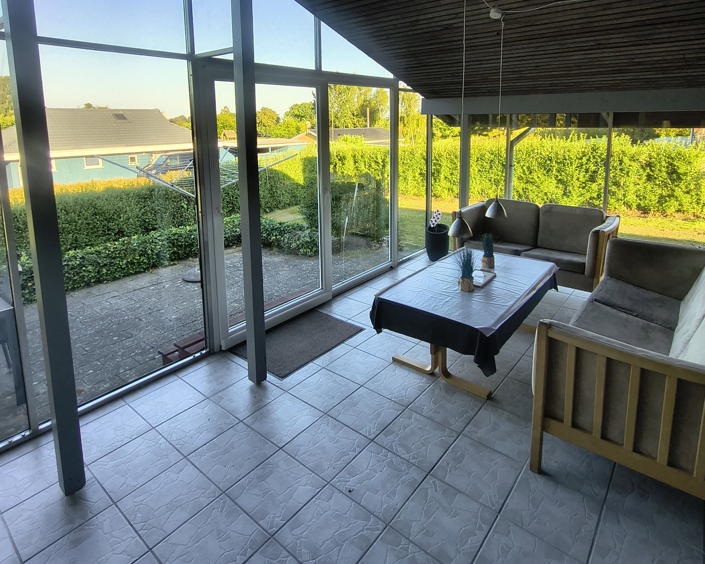 Un espacio de estar al aire libre techado con mesa y sillas, ventanas amplias y vistas a un jardín y edificios.
