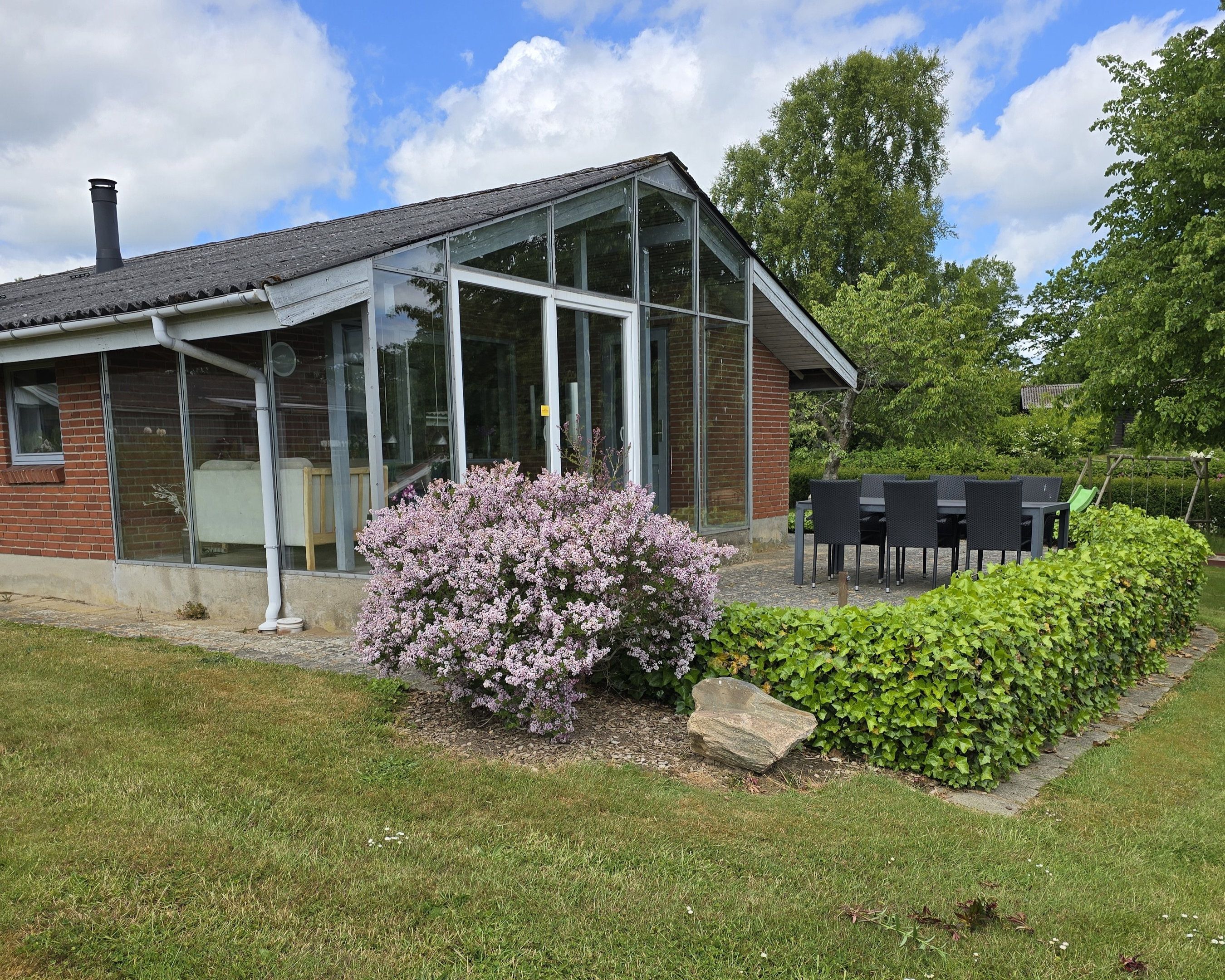 A brick building with large windows and a glass extension, surrounded by a well-maintained lawn and flowering bushes.