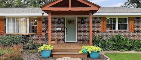 A two-story brick building with a covered porch, a front door, and a small garden area with potted plants.